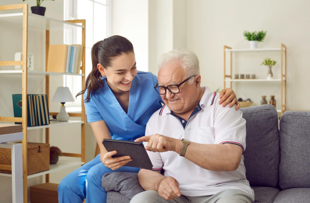 An older adult with early-stage dementia laughing beside a smiling caregiver while showing them something on a tablet.