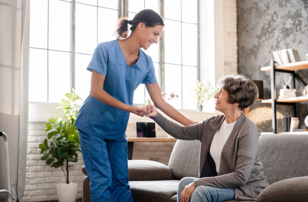 A caregiver helps an appreciative older adult stand up from the couch in front of a bright sunlit window in an assisted living apartment