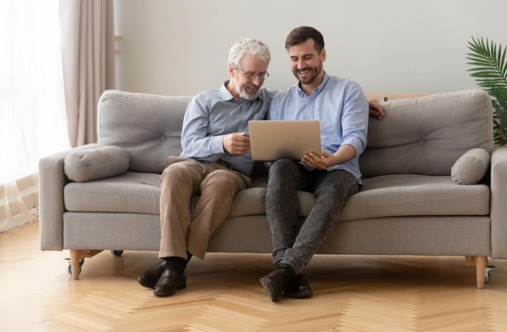 An older adult and their adult child laugh while sitting on a couch and using a laptop to research assisted living communities near them